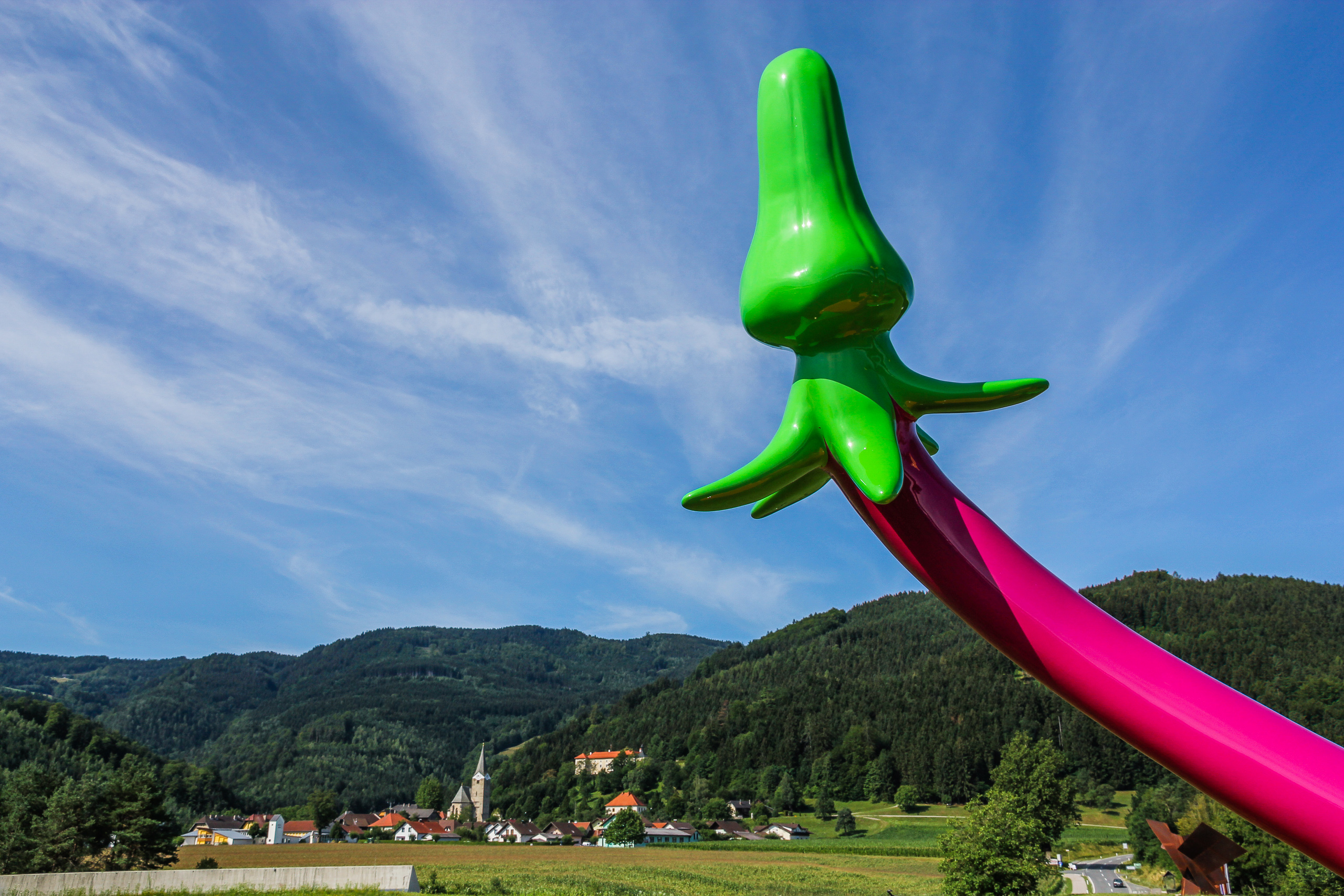Grellgrün und Pink gegen den Sommerhimmel, tief unten der Ort mit Kirche